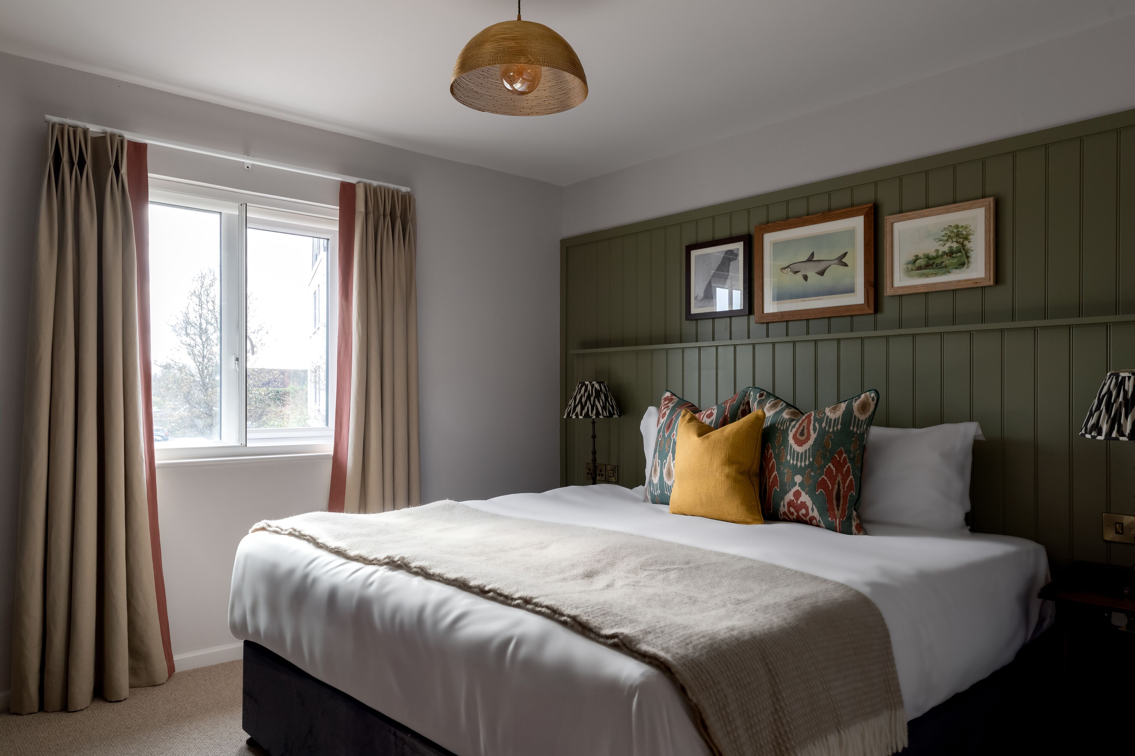 A bedroom with a large bed, a window with curtains, a hanging lamp, and framed pictures on the wall.