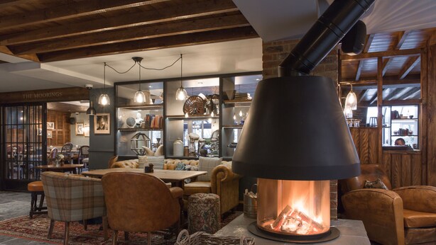 Un interior acogedor con una chimenea grande, vigas de madera y una zona de comedor con sillas y mesas.