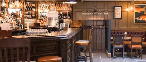 Zona de bar con taburetes de madera, un hielero grande y una variedad de botellas y vasos en estantes.
