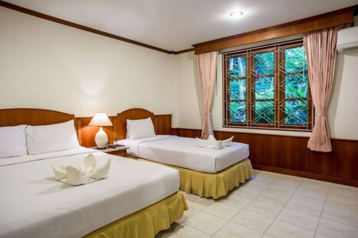 A hotel room with two beds, a nightstand with a lamp, a window with curtains, and a view of greenery outside.