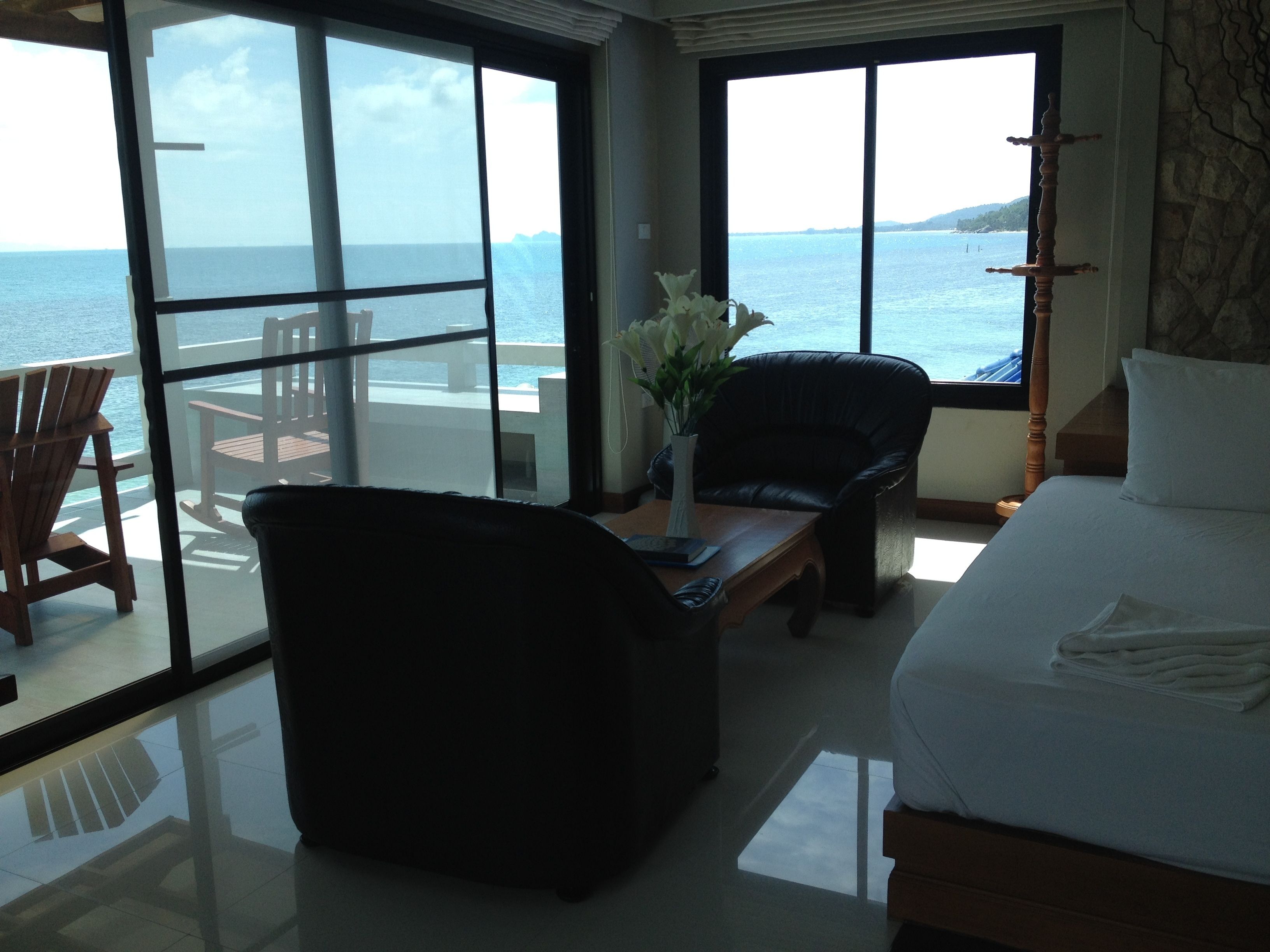 Una habitación con una cama, un sillón de cuero negro, una mesa de madera y un jarrón con flores. Hay un ventanal grande que ofrece vistas al mar y una terraza exterior.