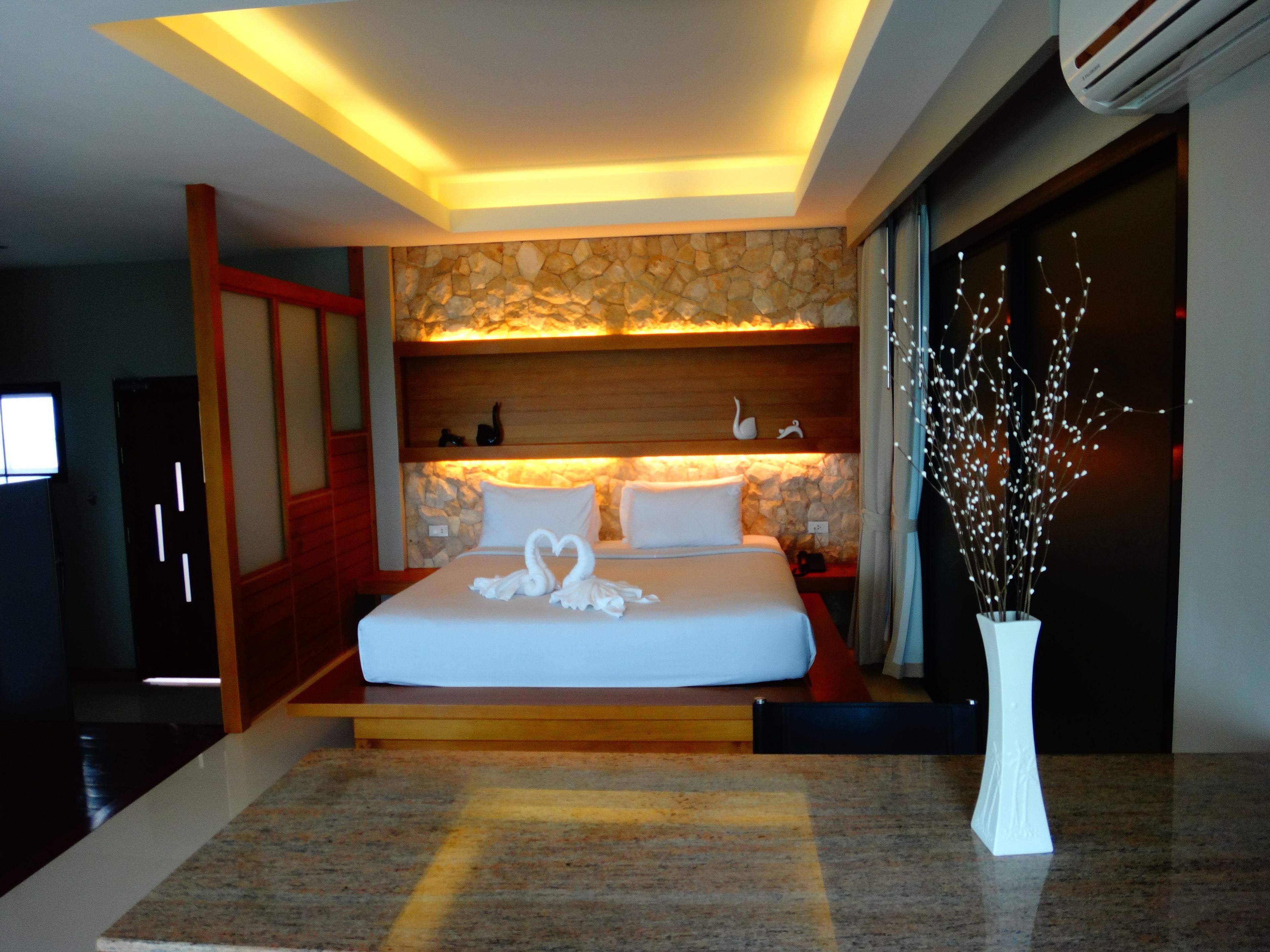 A modern hotel room with a large bed, stone wall, wooden paneling, and a vase with white flowers.