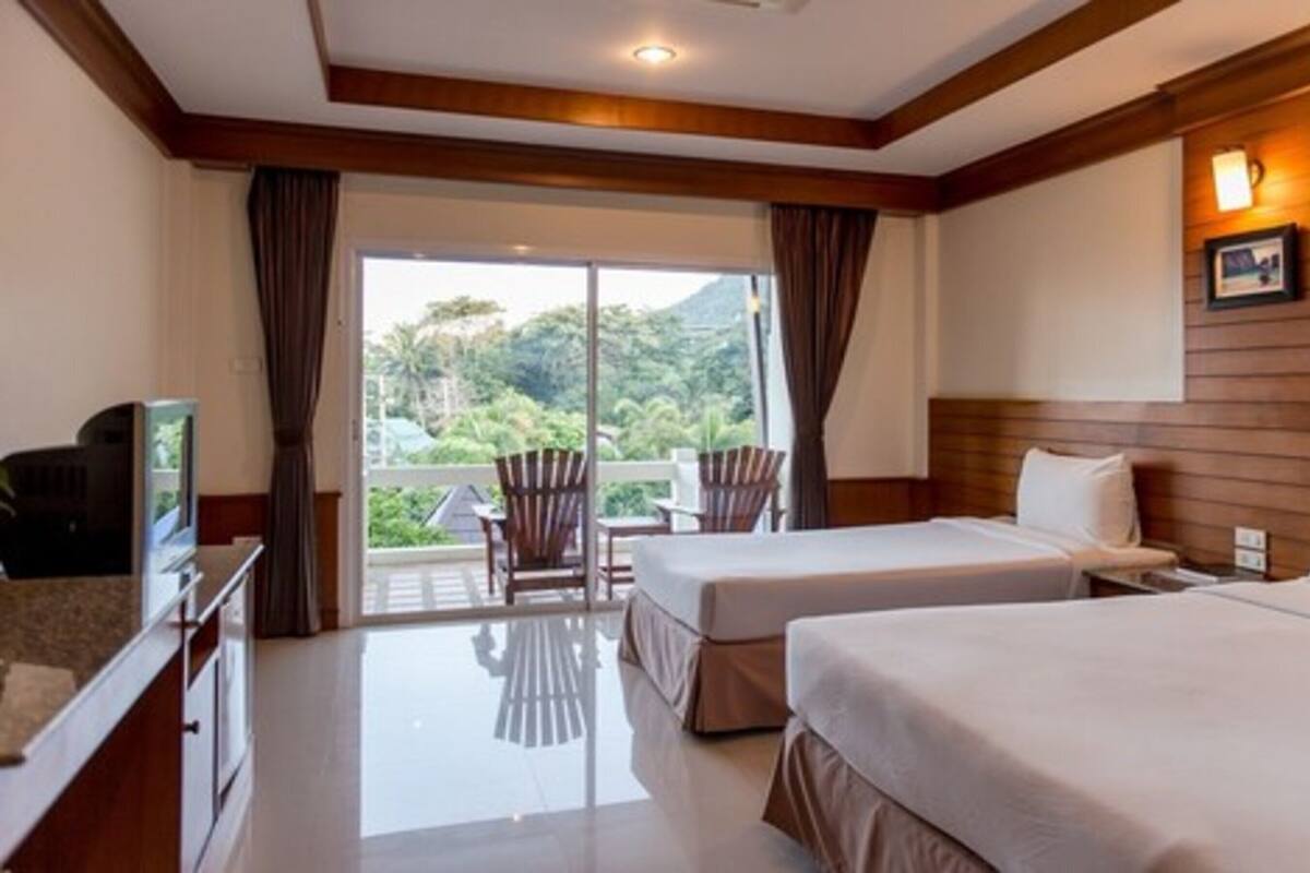 A hotel room with two beds, a TV, a desk, and a balcony with a view of greenery.