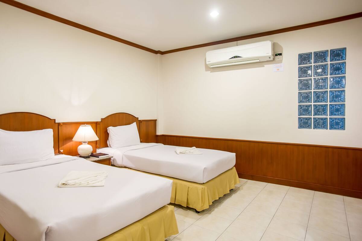 A hotel room with two beds, a nightstand with a lamp, an air conditioning unit, and a wall with a blue glass art piece.