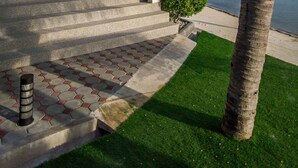 Un edificio moderno con balcón que da a una masa de agua, escalones que conducen a la entrada y una palmera cerca.
