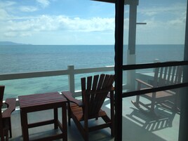 Un balcón con sillas y una mesa de madera, con vistas a un mar tranquilo y a tierra distante.