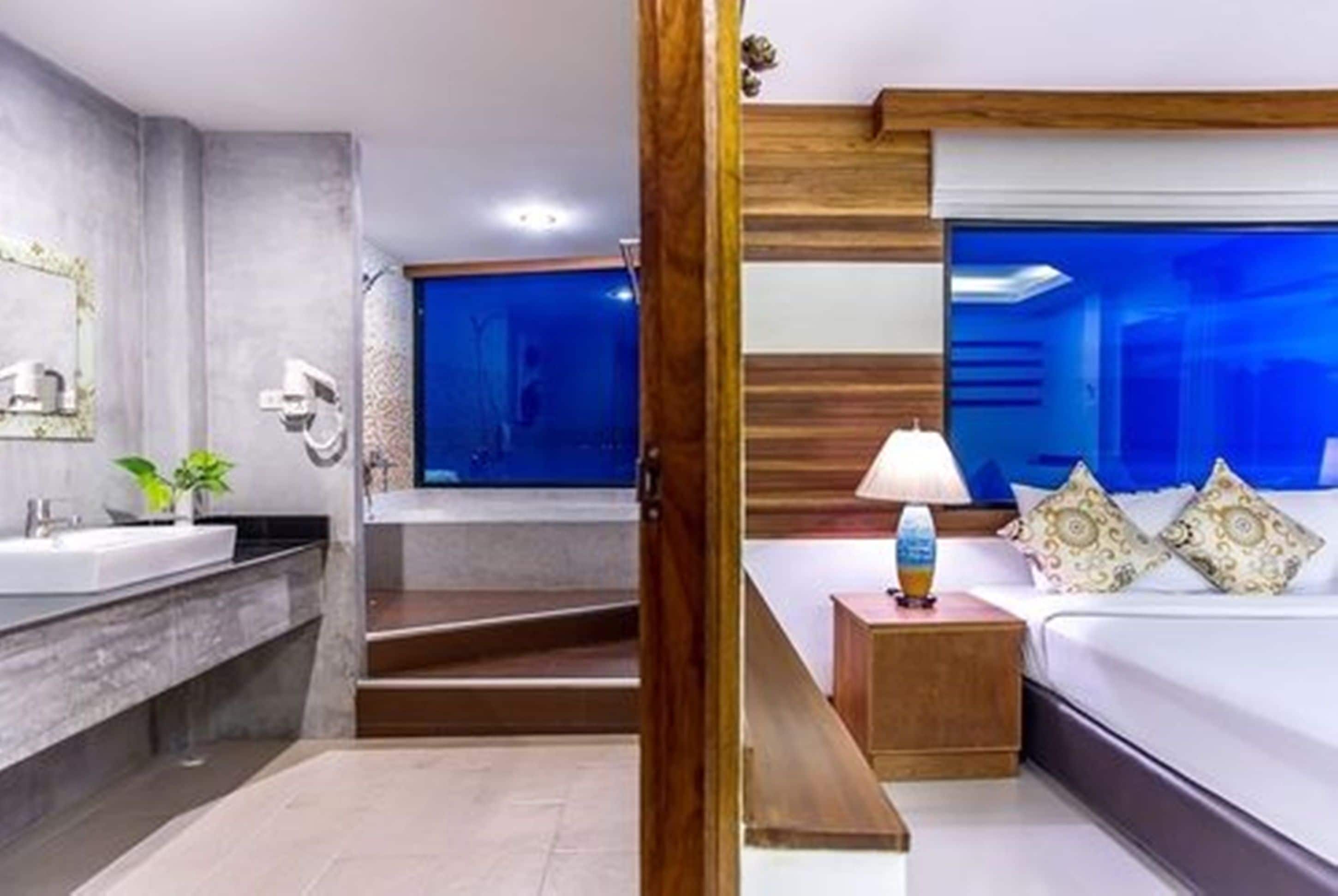 A modern hotel room with a large bed, a bedside table with a lamp, a bathroom with a large mirror, and a view of the ocean.