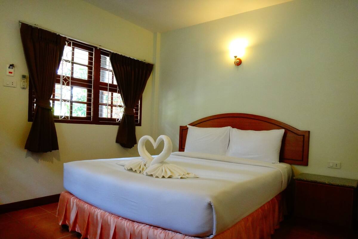 A neatly made bed with a white bedspread and a pink skirted bed skirt. There is a window with brown curtains and a wall-mounted light fixture.