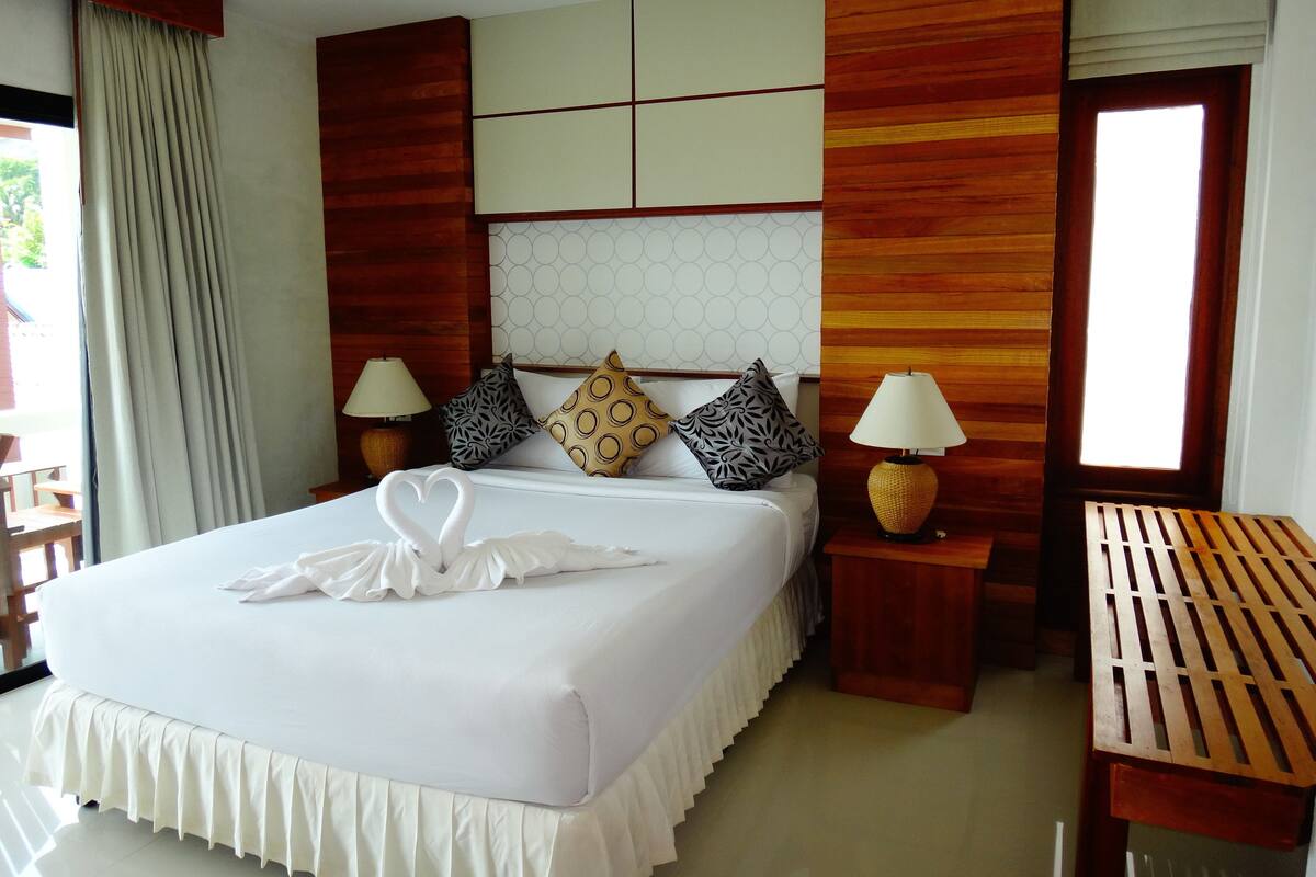 A neatly made bed with a heart-shaped towel arrangement, a wooden headboard, patterned pillows, two bedside lamps, and a wooden nightstand.