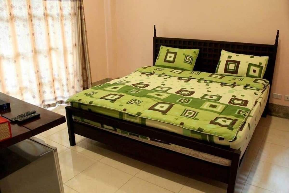 A neatly made bed with a patterned bedspread and pillows, a wooden headboard, a small table with a remote control, and a window with curtains.