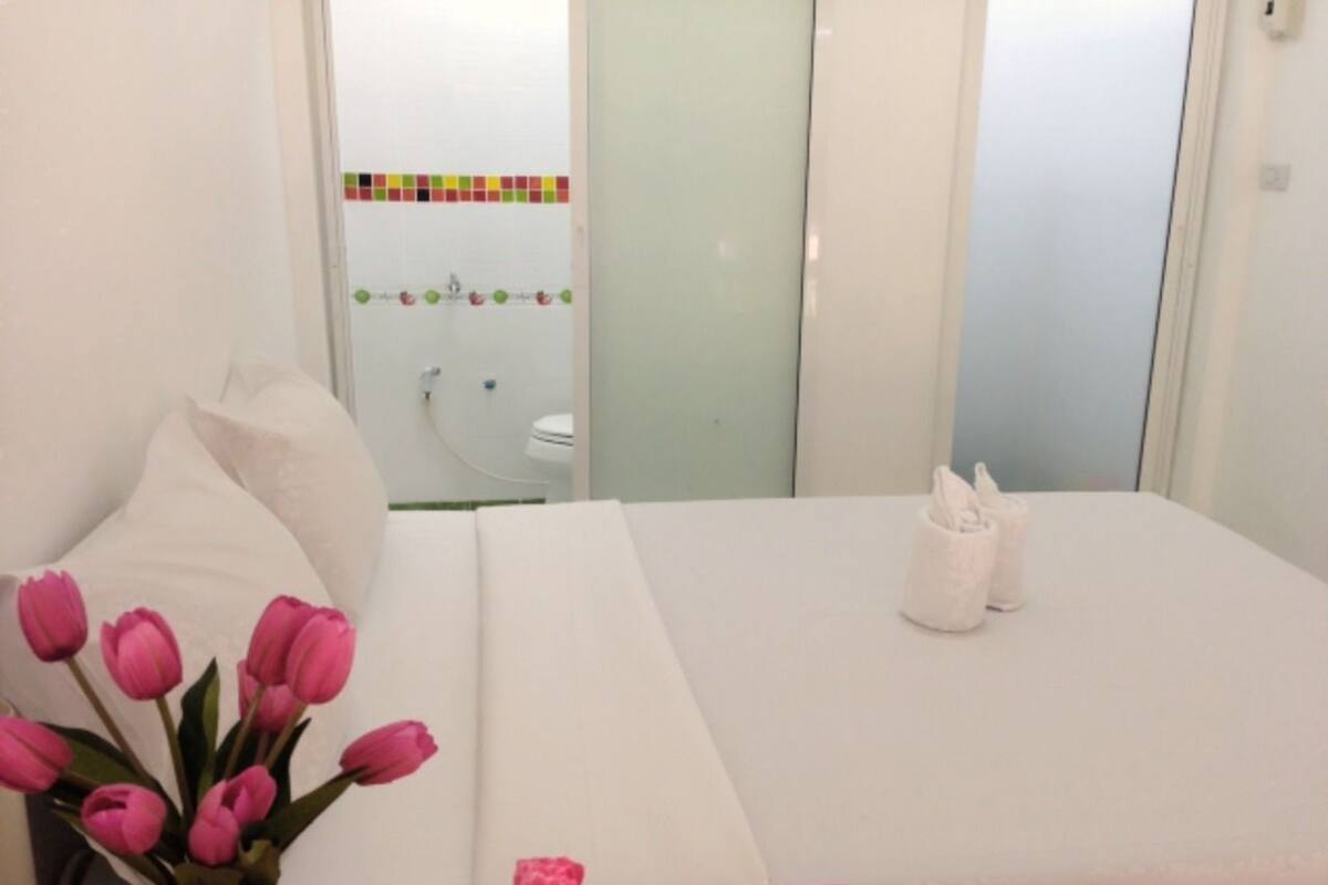 A hotel room with a bed, a bedside table with a vase of pink flowers, and a bathroom visible through an open door.
