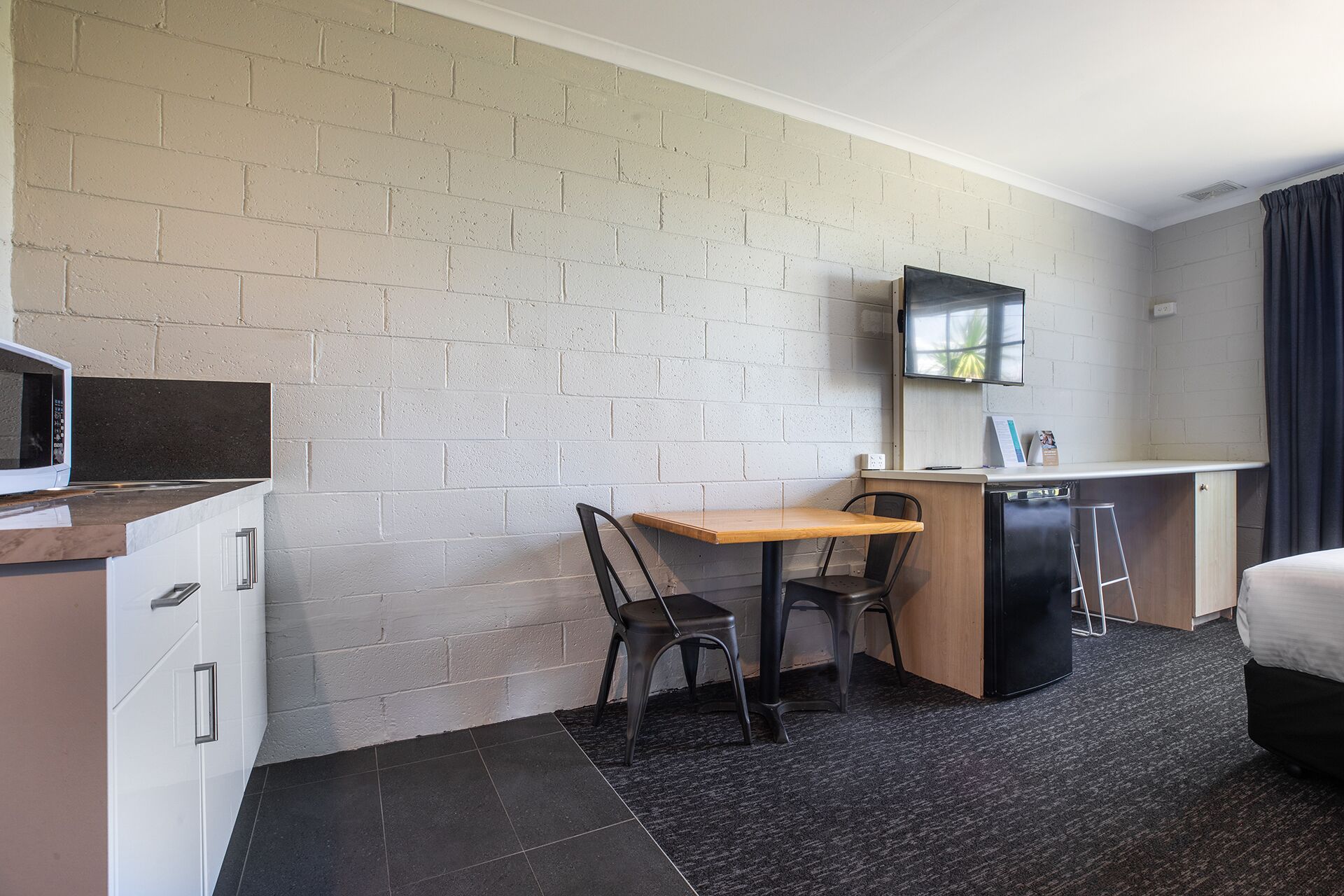 Habitación de hotel compacta con kitchenette, una cama individual, un escritorio con silla y un televisor montado en la pared.