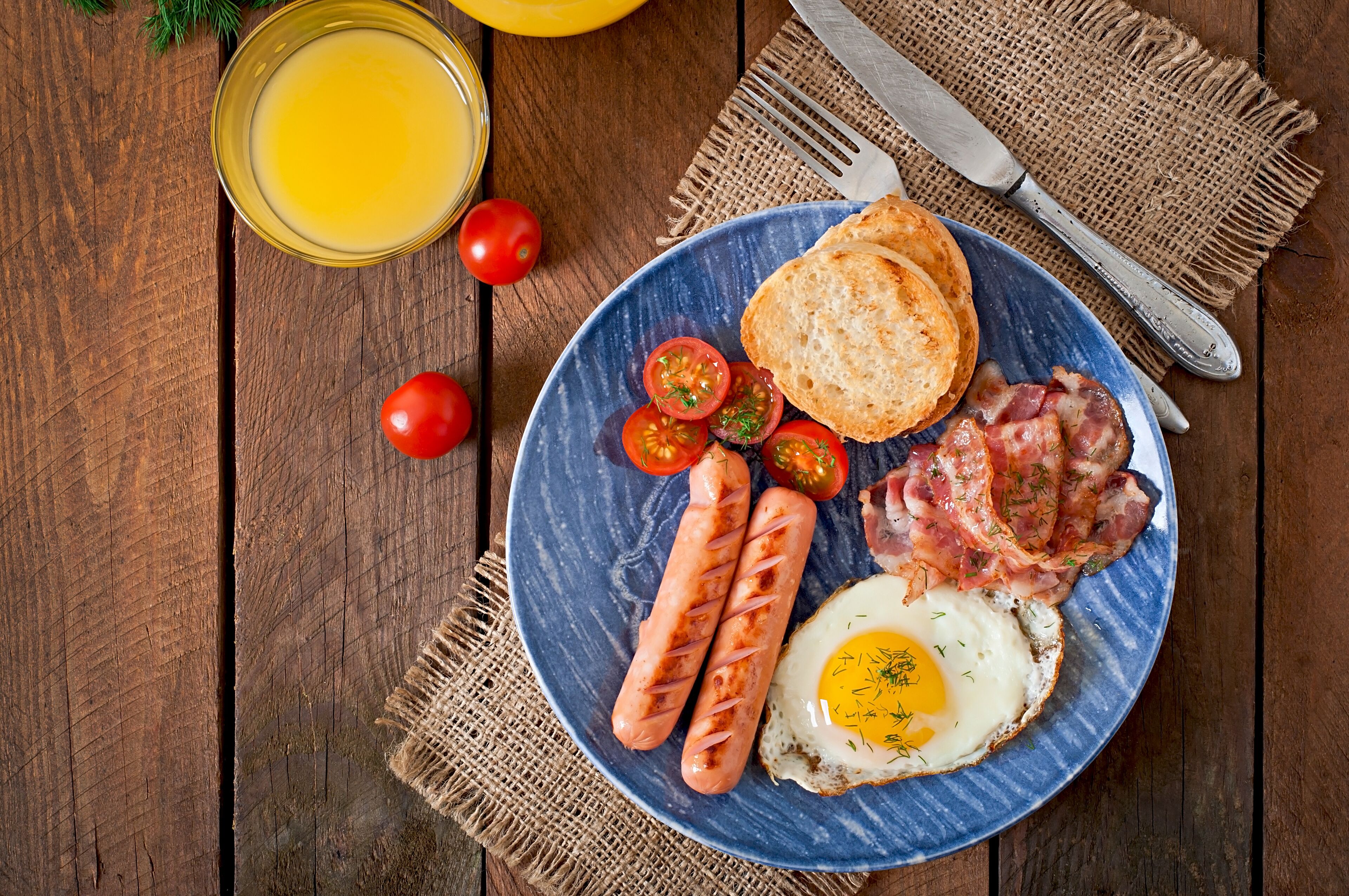 Un plato de desayuno con salchichas, tocino, un huevo frito y tomates sobre un plato azul.