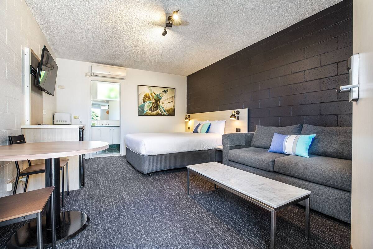 A compact hotel room with a bed, a sofa, a small table, and a TV mounted on the wall.