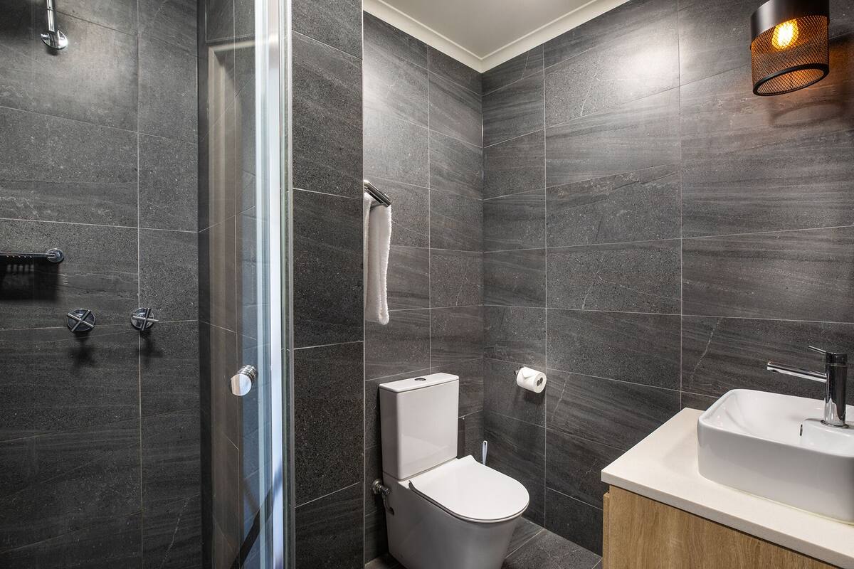 A modern bathroom with dark tiles, a glass shower enclosure, a toilet, and a sink with a wooden vanity.