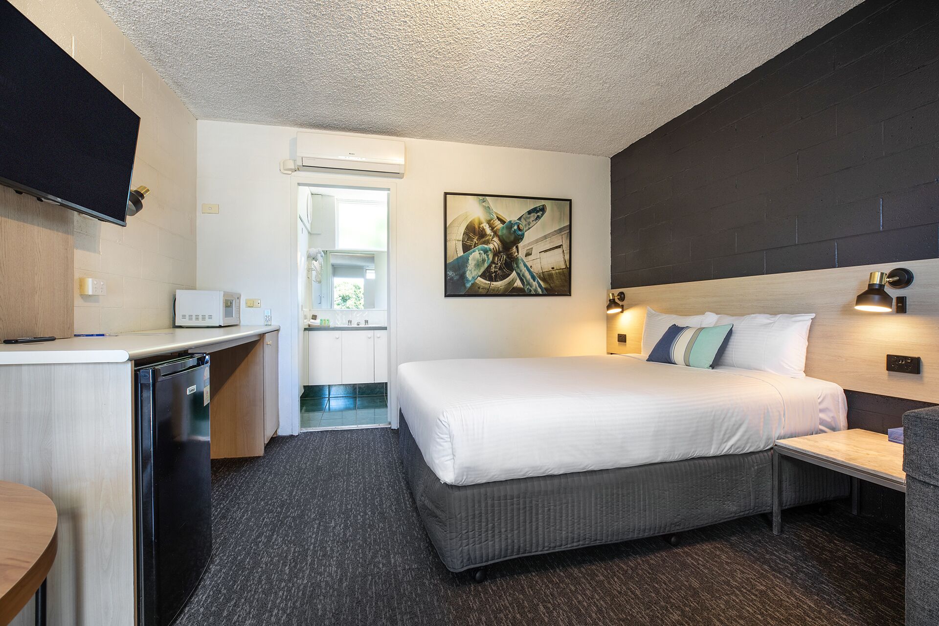 Habitación de hotel con cama, mesita de noche y un pequeño escritorio con silla. Se ve una ventana con vistas al exterior.