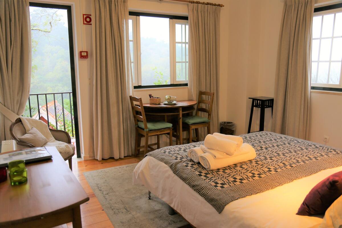 Quarto com cama, escrivaninha e cadeira. Janela com cortinas oferece vista para o verde lá fora, e uma mesinha com uma cesta.