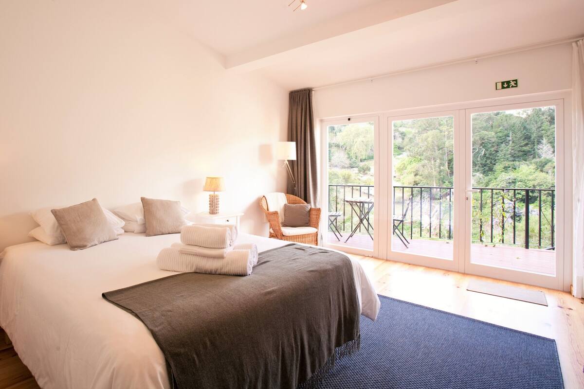 Quarto de hotel com uma cama grande, duas mesas de cabeceira com luminárias, uma cadeira e varanda com vista para áreas verdes.