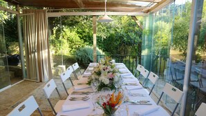 Un comedor con una mesa larga dispuesta para una comida, rodeada de sillas, y un cerramiento de cristal con vistas a la vegetación.