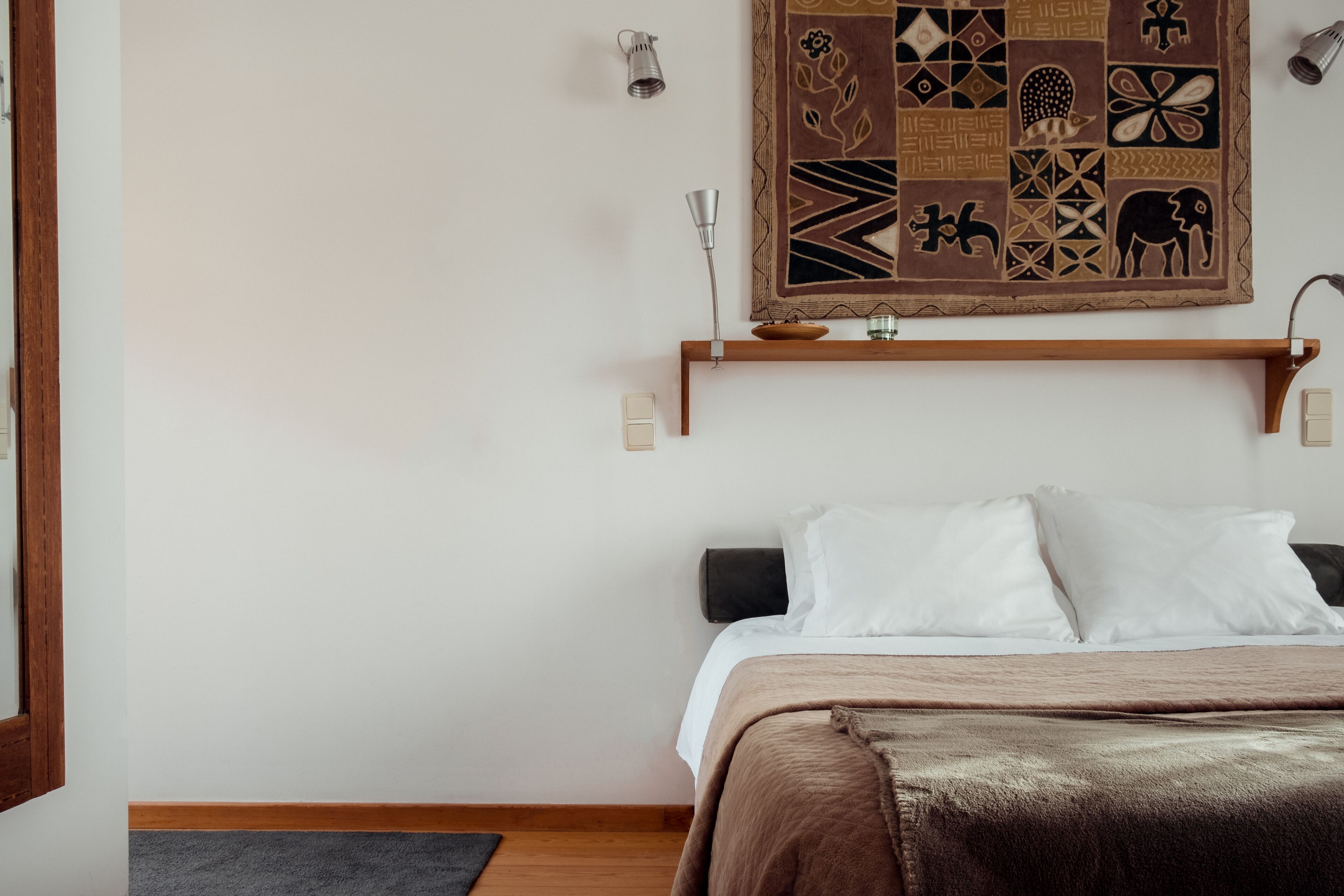 A bedroom with a bed, a wooden headboard, a wall-mounted shelf with decorative items, and a patterned tapestry.