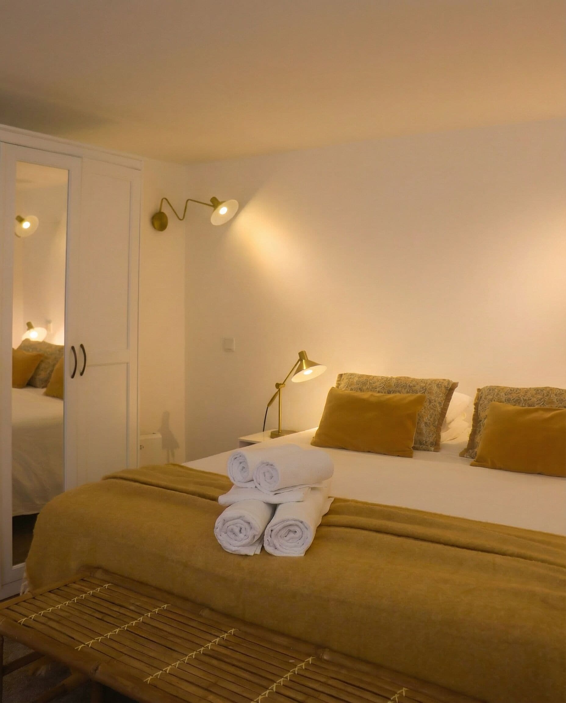 Une chambre d’hôtel avec un lit, une table de chevet avec une lampe, des serviettes pliées sur le lit et un banc au pied du lit.