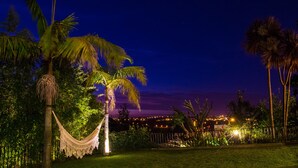 Vista nocturna de un jardín tropical con una hamaca, palmeras y un paisaje urbano lejano.