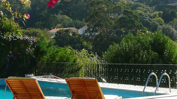 Zona de piscina con dos sillones de madera y vista a una colina con vegetación y edificios.