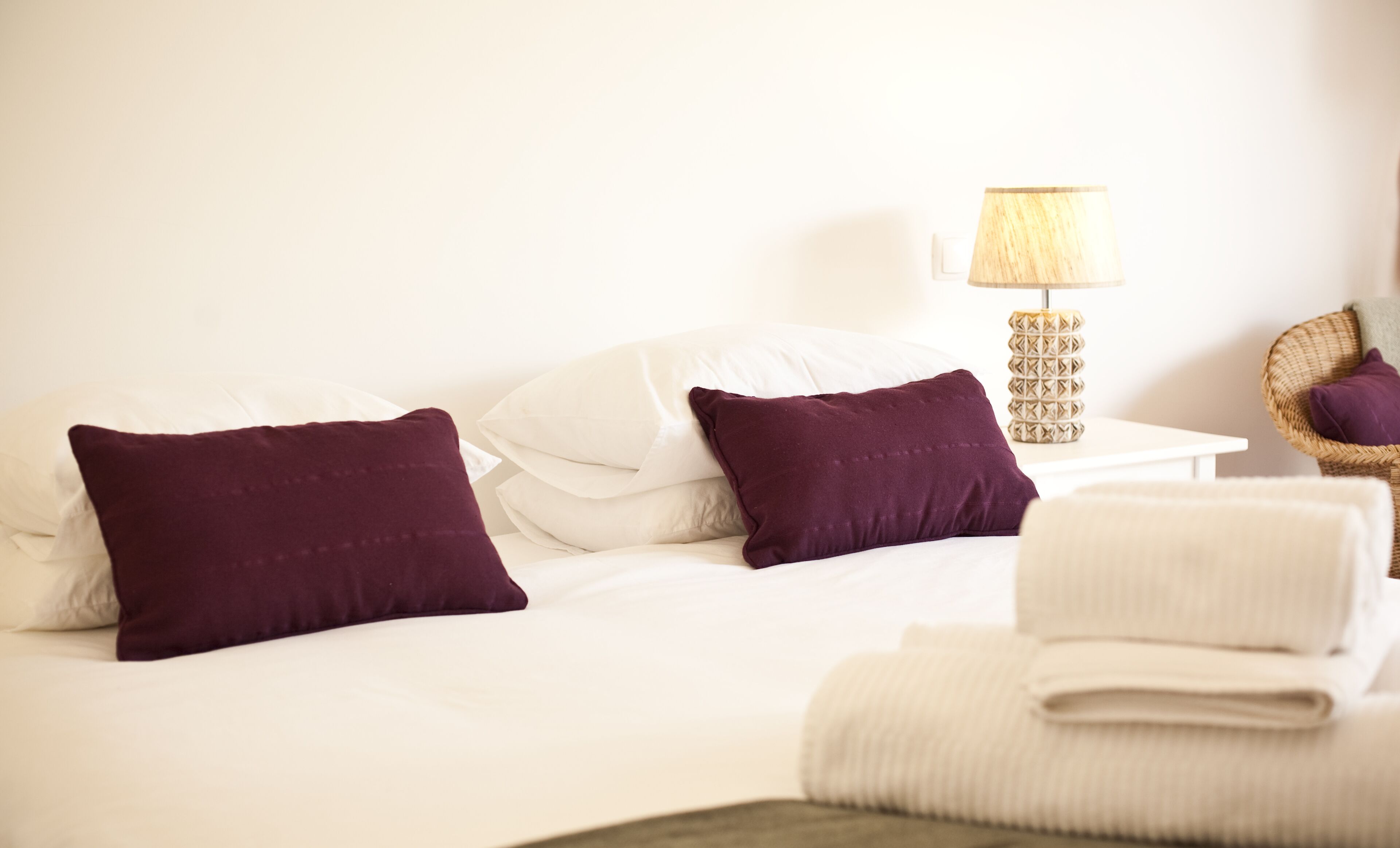 A neatly made bed with white and purple pillows, a small bedside table with a lamp, and a wicker chair with a cushion.