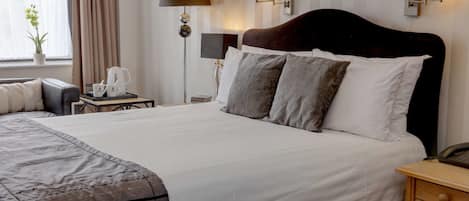 Een net opgemaakt bed met een donker hoofdbord, een nachtkastje met een lamp, en een klein tafeltje met een koffiezetapparaat en kopjes.