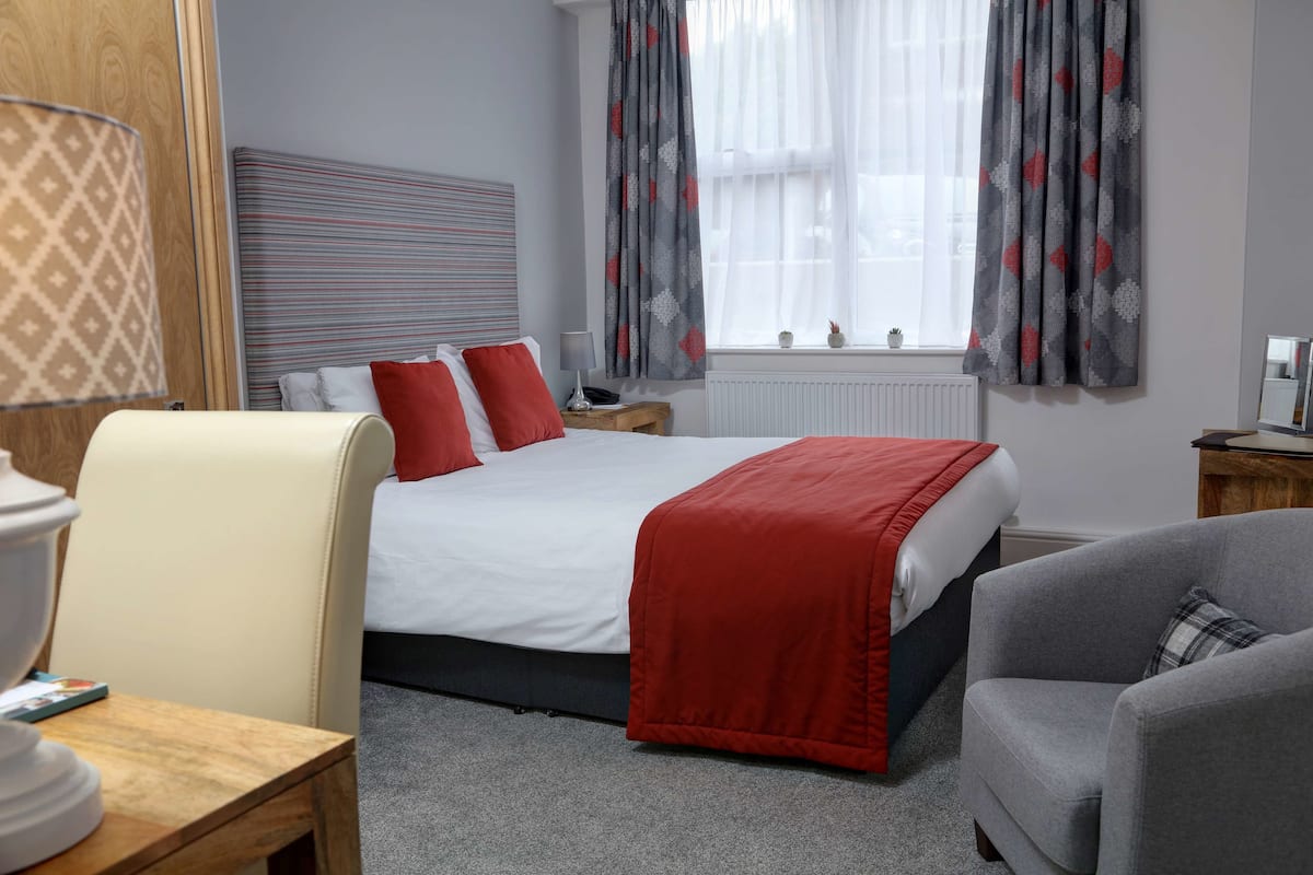 Une chambre d’hôtel avec un lit, deux tables de chevet, une lampe, une chaise et une fenêtre avec des rideaux.