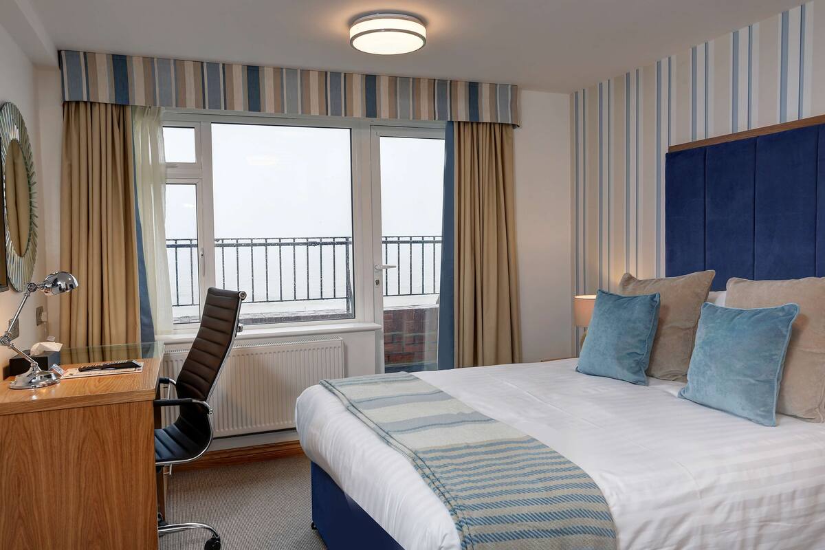 Une chambre d’hôtel comprenant un lit, un bureau, une chaise, une fenêtre avec des rideaux et donnant sur le balcon extérieur.
