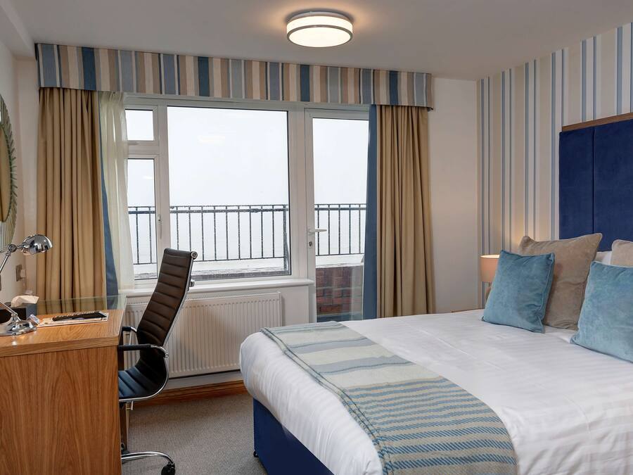 A hotel room with a bed, a desk, a chair, a window with curtains, and a view of the outside balcony.