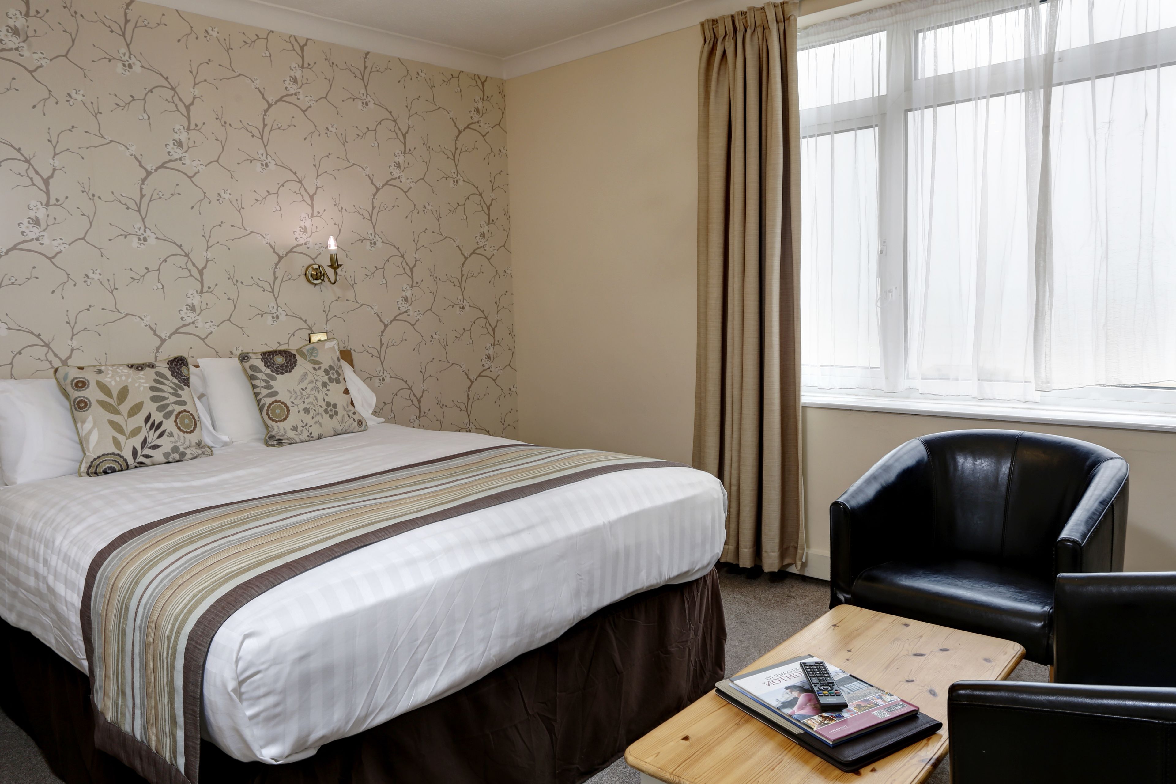 A hotel room with a bed, a wooden coffee table, a black leather chair, and a window with curtains.