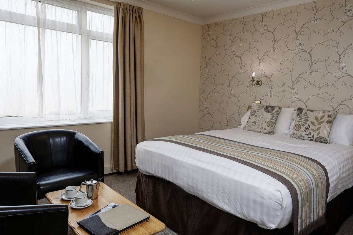 Une chambre d’hôtel avec un lit, une chaise, une table sur laquelle se trouve un plateau avec des tasses et un carnet, ainsi qu’une fenêtre avec des rideaux.