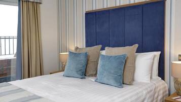 A neatly made bed with a blue headboard and beige and blue pillows, a striped curtain, and a window with a view of a balcony.