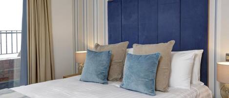 Een netjes opgemaakt bed met een blauw hoofdeinde en beige en blauwe kussens, een gestreept gordijn en een raam met uitzicht op een balkon.