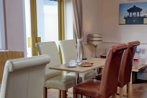 Un coin repas avec une table mise pour deux, agrémenté d’un pichet blanc, d’une tasse et d’une assiette de nourriture. Il y a deux chaises, une blanche et une marron. Une lampe et un tableau représentant un belvédère sont accrochés au mur.