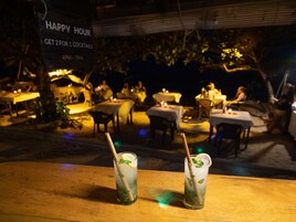 Escena nocturna en un restaurante al aire libre con mesas y sillas, y dos vasos de una bebida refrescante a la menta sobre una mesa de madera.