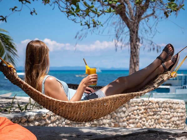 A person relaxing in a hammock by the sea, holding a drink.