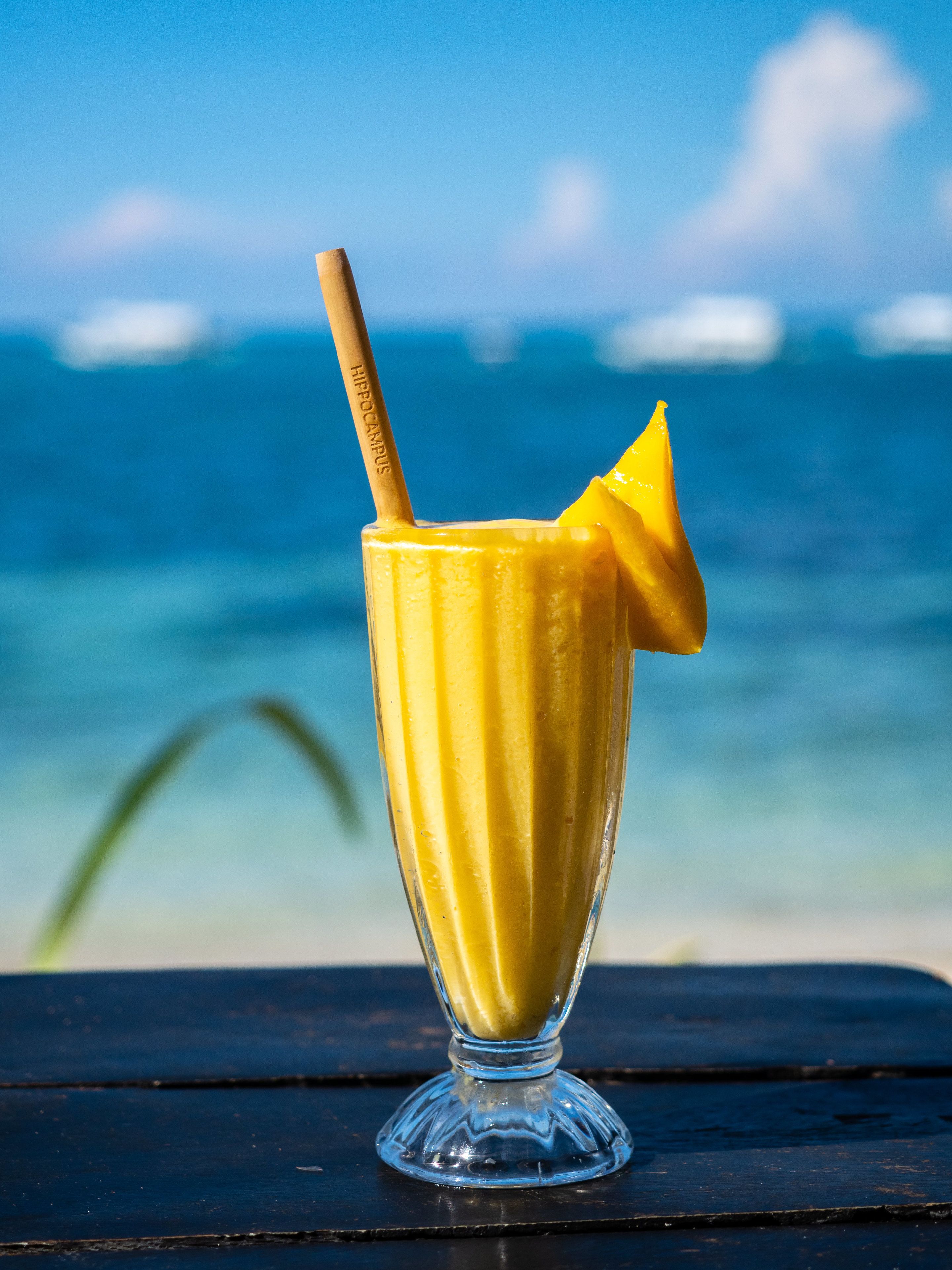 A glass of yellow drink with a straw and a slice of fruit on a table by the sea.