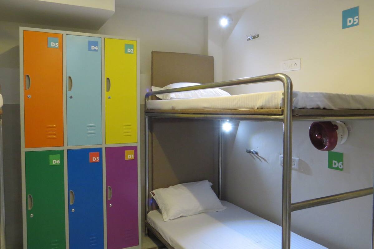 A bunk bed with a white mattress and pillow, a metal frame, and a wall-mounted fan. Adjacent to the bunk bed is a set of colorful lockers with labels D1 through D6.