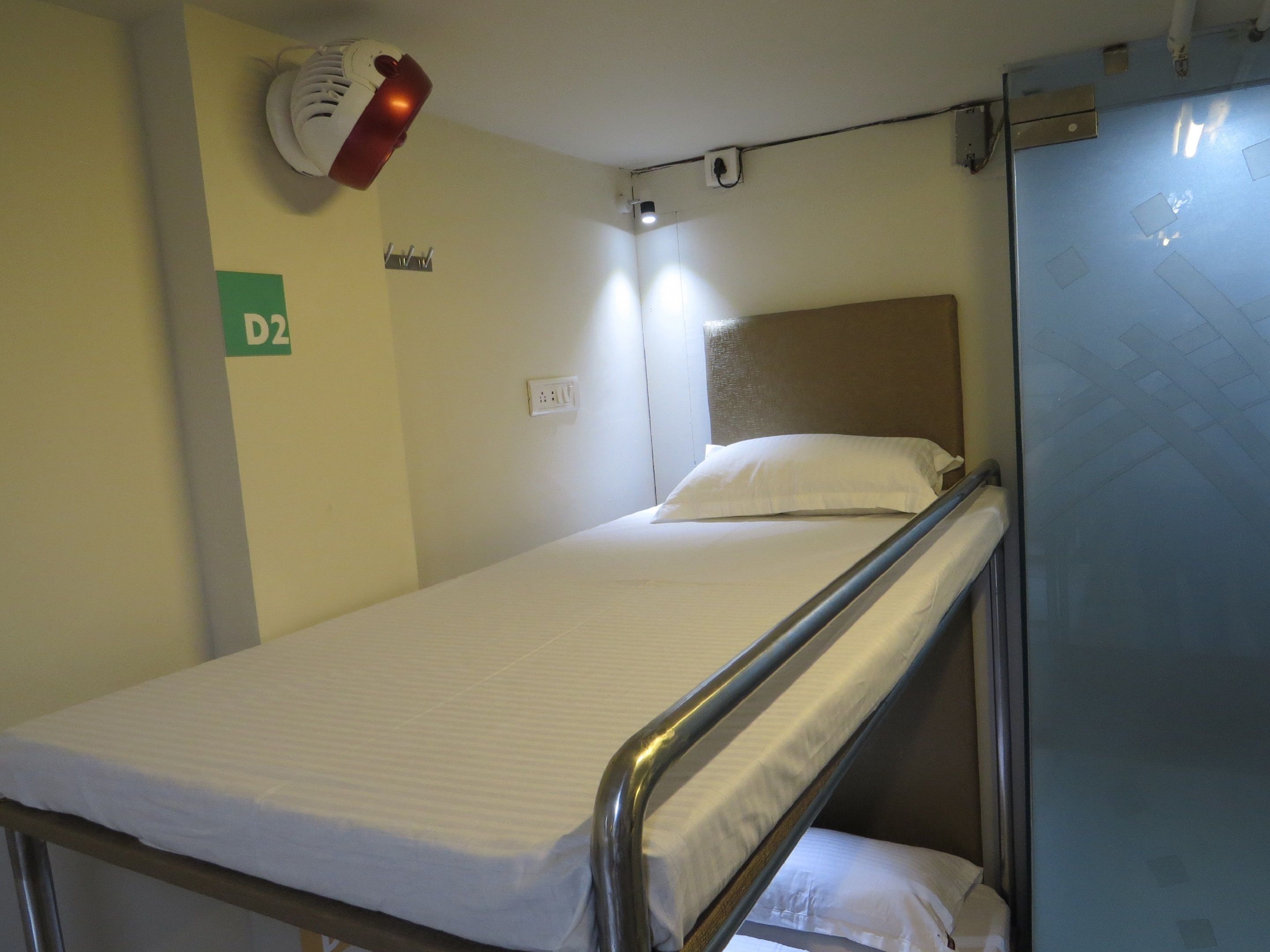 A bunk bed with a metal frame, a white mattress, and a headboard. There is a wall-mounted light fixture above the bed and a small shelf with a hook.