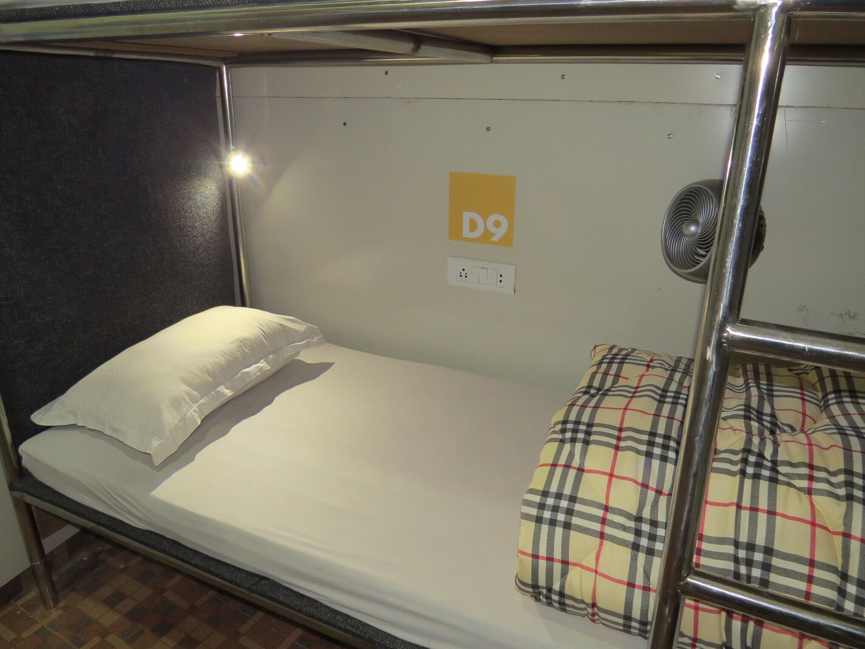 Un lit superposé avec un matelas blanc et une couverture à carreaux. Un petit panneau jaune portant la lettre « D9 » est accroché au mur.