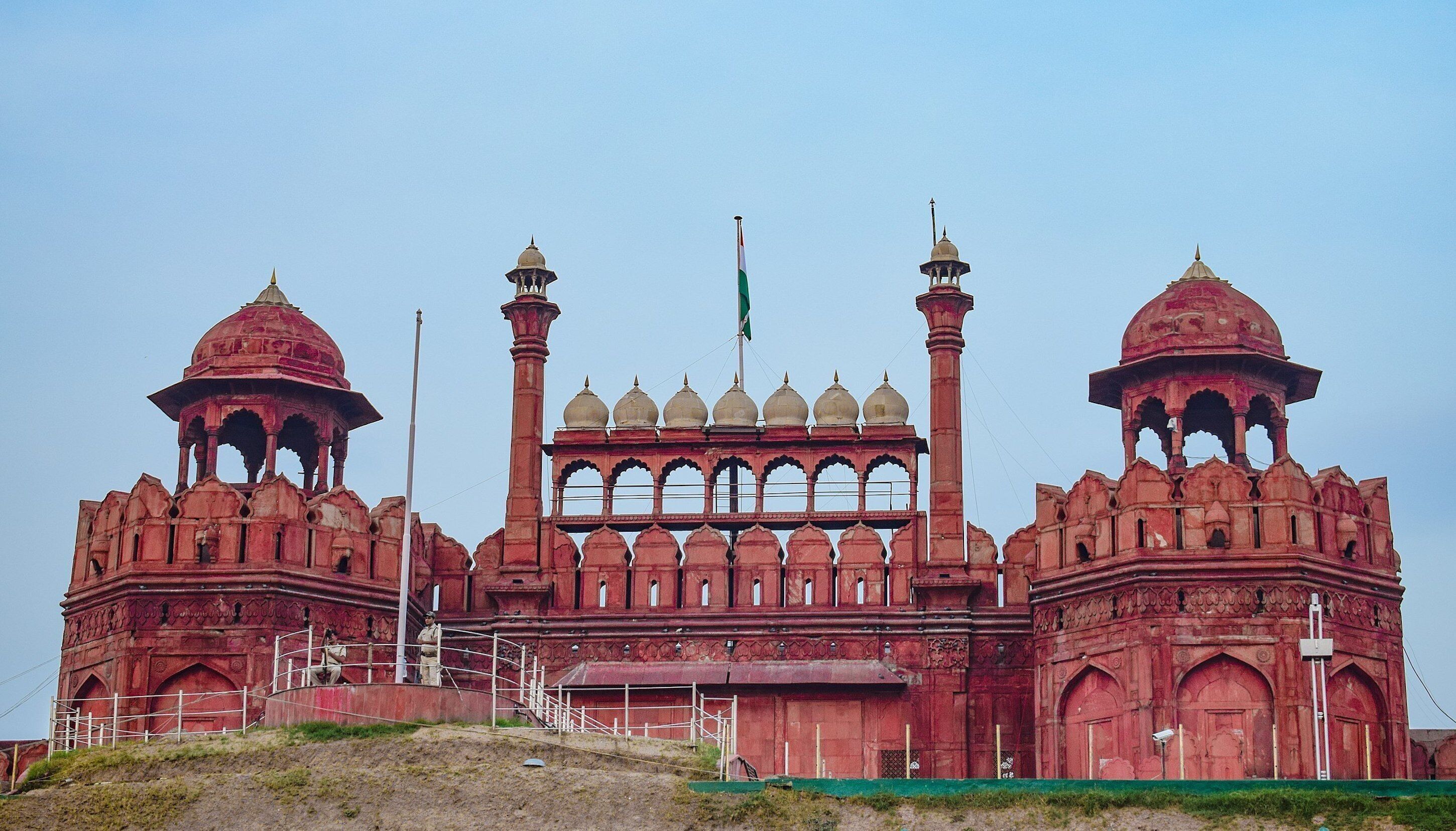 A large, red, historic building with multiple domes and arches, featuring a prominent flagpole.