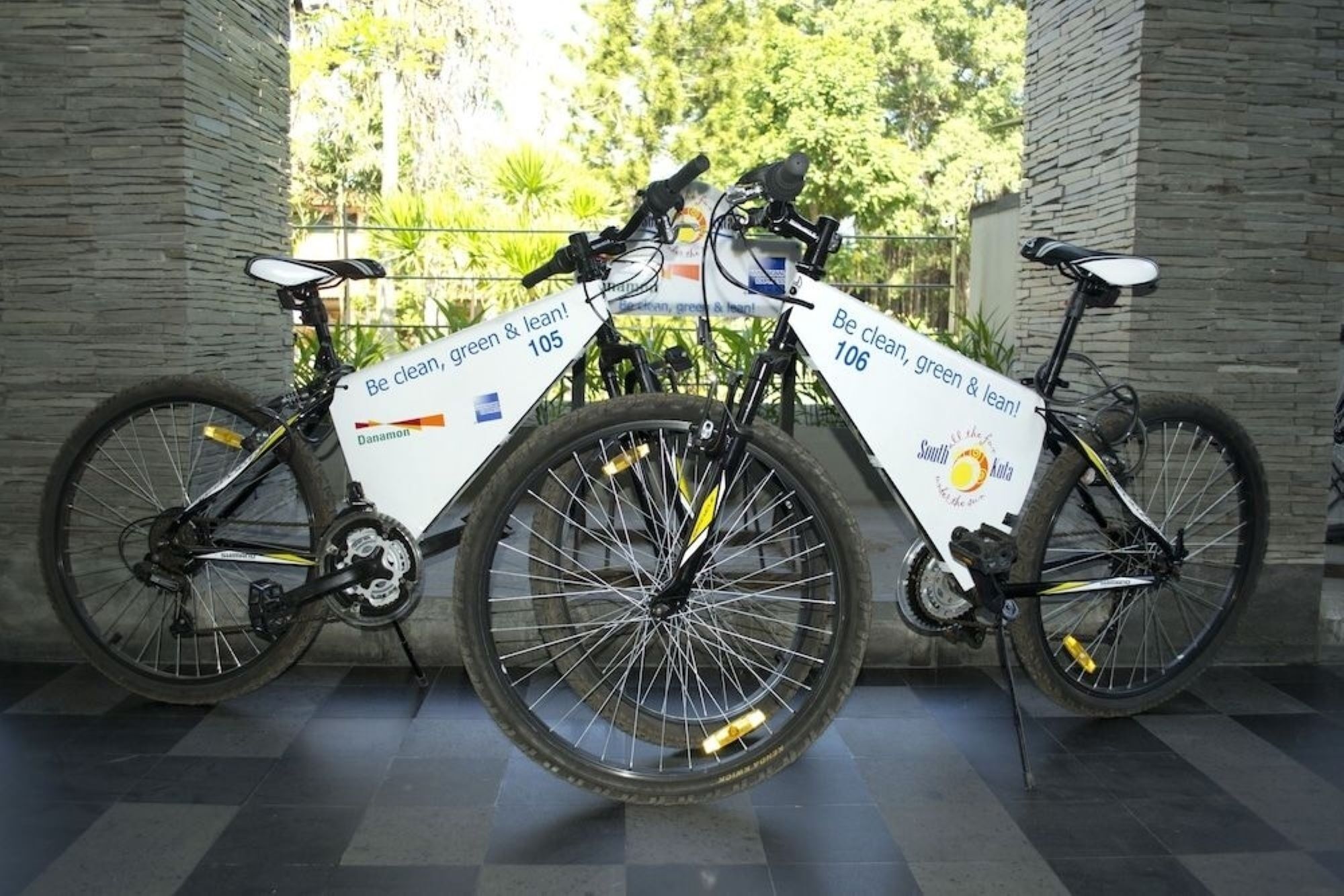 Zwei Fahrräder mit weißen Bannern, eines mit der Aufschrift '105' und das andere mit '106', in einem überdachten Bereich mit gefliestem Boden und einer Steinwand abgestellt.