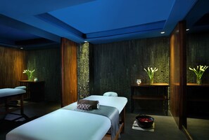 A spa room with two beds, a massage table, and a wall with vertical wooden slats. - Bali Dynasty Resort (Kuta)
