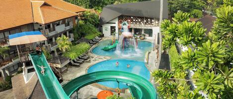 A water park with multiple slides and pools, surrounded by lush greenery and a building with a tiled roof.