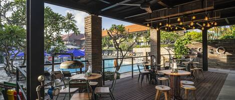 An outdoor restaurant with wooden tables and chairs, a pool in the background, and a view of the surrounding area.
