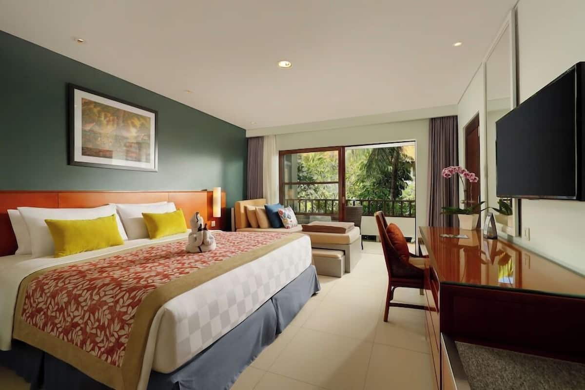 Hotelkamer met een bed, bureau, stoel, televisie en een groot raam met uitzicht op groen.