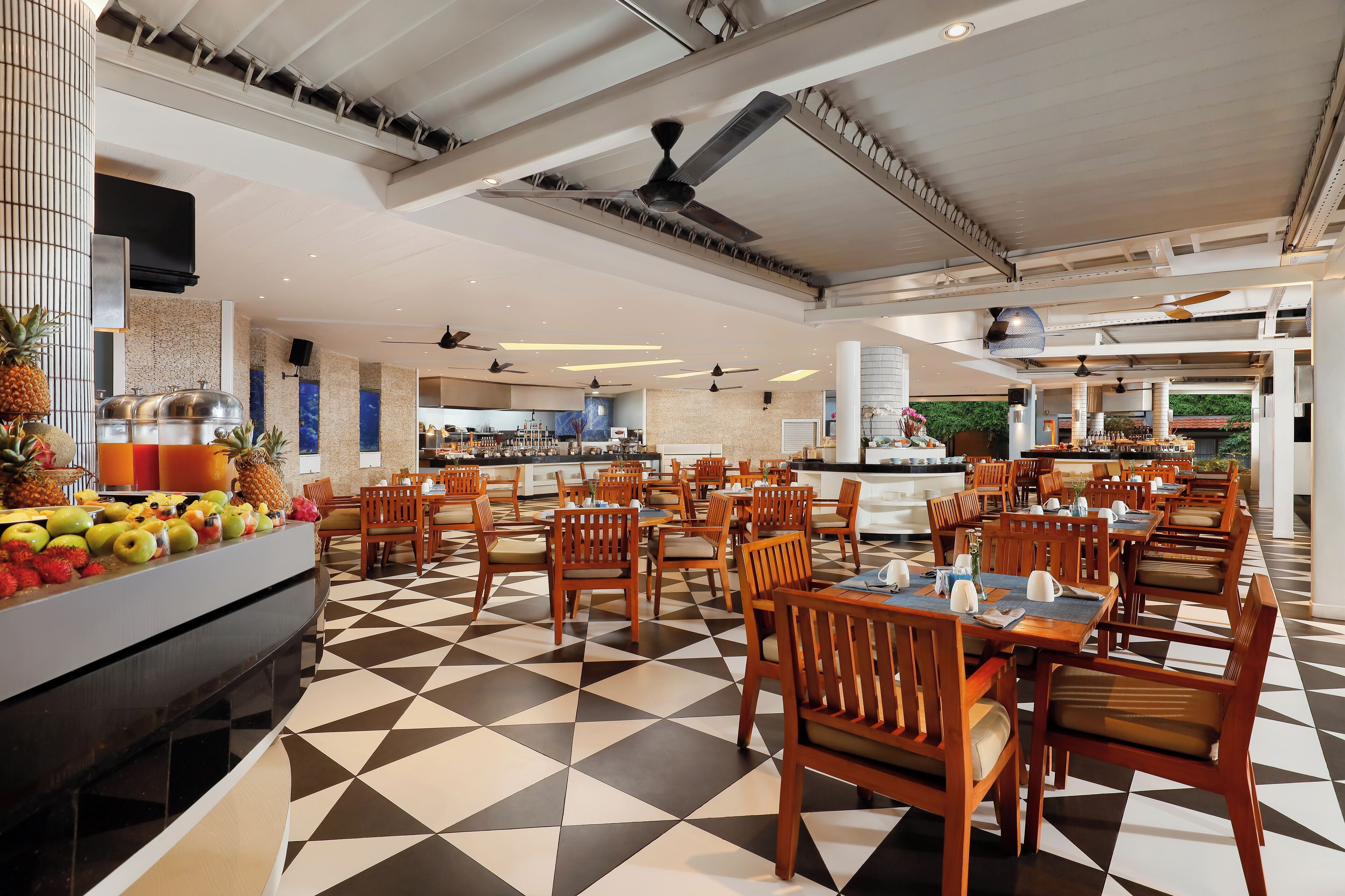 A hotel dining area with a checkered floor, wooden chairs, and a buffet with fruits.